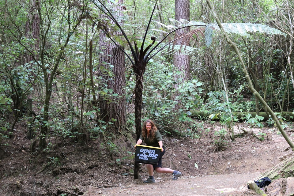 A woman with long curly light brown hair holding a large plastic container under a punga tree on a dusty dirt track in amongst tall native forest.