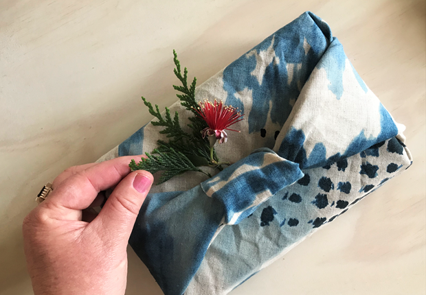 A hand placing a pohutakawa blossom into the bow on a gift which has been wrapped in hand dyed blue fabric.