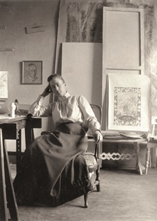 A woman in Victorian clothing, a white shirt and long dark skirt, sitting on a chair and her elbow on the table with head resting on her hand, with an easel and artwork leaning up against a wall behind her. Image is black and white.