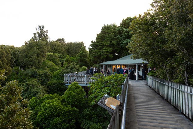 A long boardwalk leading to a building, lit from the inside and surrounded in people, set in the canopy of native bush.