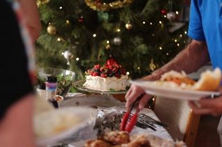 A decorated Christmas tree with table in foreground topped with pavlova and plates of food and people serving it onto their plates.