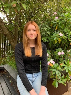 A teenage girl with long strawberry blond hair, wearing a black long-sleeved top and light blue jeans, perched on a bench seat in front of a pink flowering shrub.