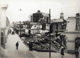 A black and white photo of a building that has been knocked down with piles of wood still lying on the site.
