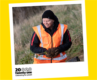 Wendi Henderson wearing a black beanie and jumper with an orange high-vis vest on top, tilting her head down towards the long grass in which she is standing and is pictured out of focus.