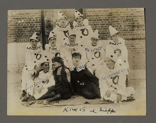 A sepia photo of a perfomance troupe wearing white pierrot costumes with two in the centre wearing black clothes and stage makeup.