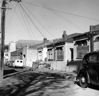 A black and white photo of Whittaker Street in 1969 where a row of small wooden houses stand and a few old motorcars are parked.
