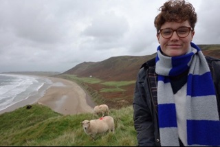 Vondy wearing a stripy scarf and smiling from the top of a cliff with a couple of sheep in the background.