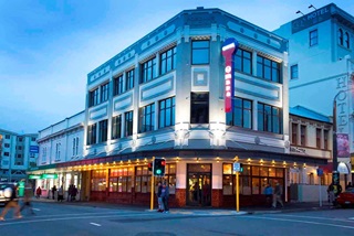 A grand old three-storey art deco building standing on a corner of two main roads intersecting, with an intricate grey and white paint job, and a blue sky behind at dusk. 