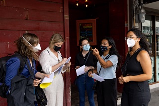 Five young women wearing masks, standing in a huddle and holding paperwork, on the street next to a maroon-coloured building.