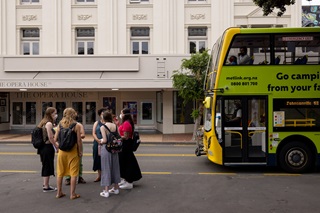 Six young women standing in a circle talking on the footpath opposite the Opera House, as a double-decker bus approaches on the road beside them.