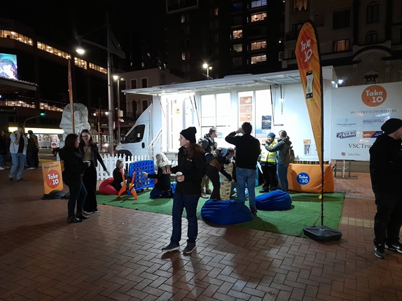 The Take 10 white caravan with green artificial turf laid out in front with blue bean bags, and people standing around on the orange pavement in the dark, with lit windows of high rises behind.