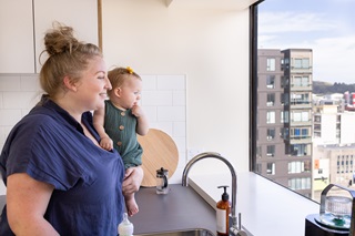 A young woman with a blond bun and blue shirt holding a toddler, standing over a modern sink, overlooking the city from a high-rise apartment window.