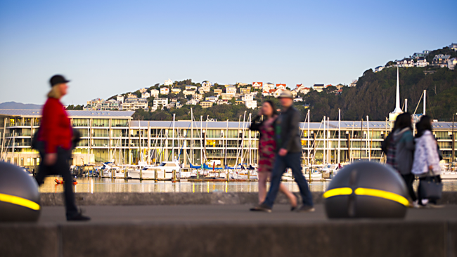 People walking along a concrete wharf in the morning light of a lovely day, with yachts, large glass terminal building, and hillside with houses in background.