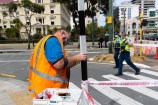 A man in a hi-vis vest removes something from a pedestrian crossing sign while two Police walk past in the background.