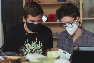 Two young people with short dark hair wearing masks looking down at a table of sewing supplies including a pin cushion.