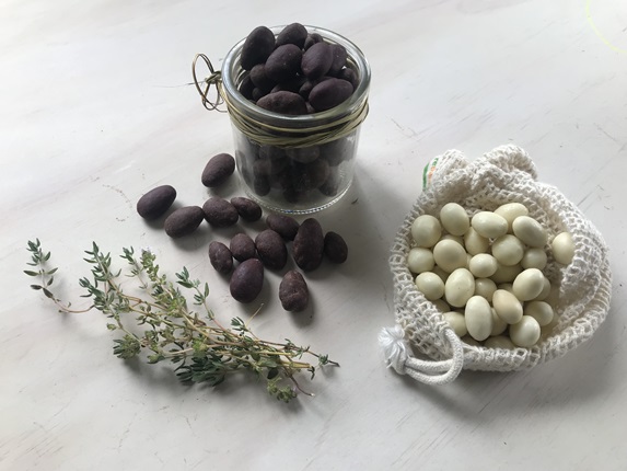 Fresh thyme, on a marble table next to a jar of black sugared almonds and a white cotton mesh drawstring bag of white sugared almonds.