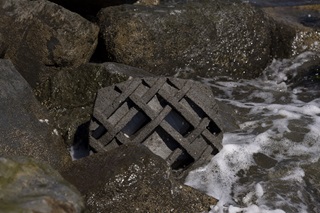 A large concrete tile with a woven pattern printed into it, placed among rocks, with the ocean washing over it.
