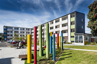 A large block of public housing flats, with a carpark to the left, and a large grassed area, wooden seating, and a bright display of coloured polls in front.