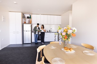 A young couple looking at a cookbook in the middle of a modern kitchen with dining table and vase of flowers in foreground.