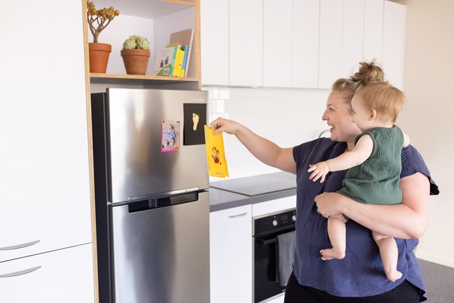 A young mum holding her baby daughter as she puts a picture on a stainless steel fridge in a white, modern kitchen.