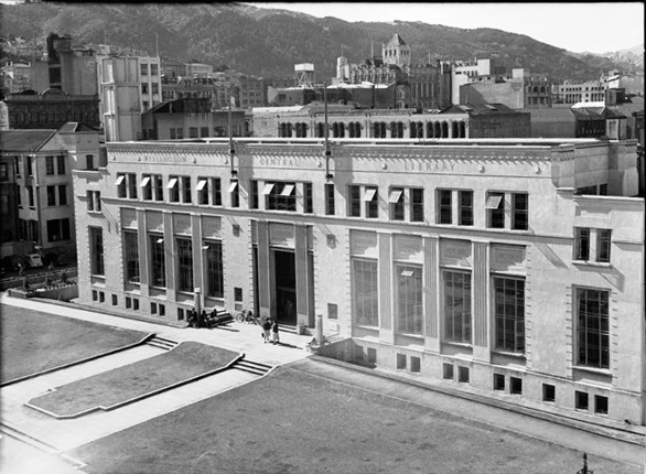 A high-angle black and white shot looking down at the old Wellington Public Library, an Art Deco building, with hills and tall old-fashioned buildings behind.