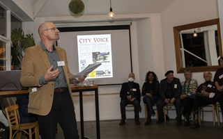 Gabor Toth presenting to a room of people in front of a projector screen which has an image of City Voice on it.