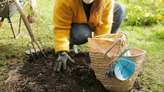 A person in jeans and a yellow knitted jumper crouching over a pile of compost being spready out on a lawn, with a woven basket and gardening fork on the side.
