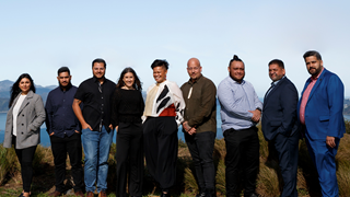 Nine relatively young people of Māori decent dressed in smart casual and standing in a line looking at the camera with clear blue sky above.