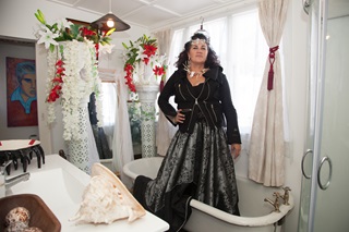 A glamourous Māori woman standing tall dressed in a glitzy long back dress standing in a white room decorated in red and white flowers.