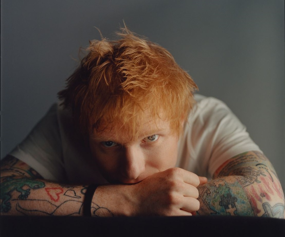 Portrait of Ed Sheeran looking at the camera while resting chin on tattooed arms on table.