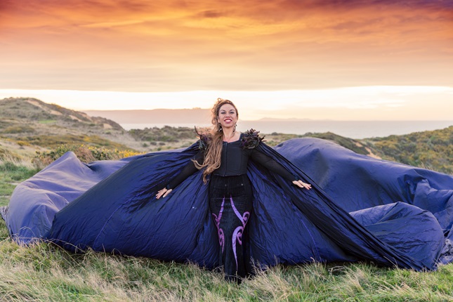 Singer Toni Huata with a billowing purple cape in a dramatic outdoor setting.
