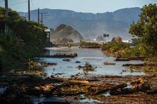 Branches and debris strewn across a flooded road. Island Bay's island is in the background, along with Wellington's eastern hills. The sky is blue after the storm has passed.