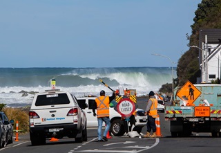 Work crews in orange high-visibility gear with their vehicles and road closed signage cordoning off a coastal road, with massive ocean swells visible beyond them, and a blue, cloudless sky.