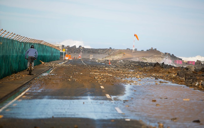 A cyclist riding over debris and rocks strewn over a wide road, which runs alongside the coast. The sky is blue following the storm.