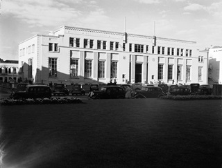 Black and white photo of a building with cars lined up infront of it.