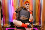A person with heavy make-up and a drawn on moustache wearing eccentric orange and black clothing, posing in a chair in front of a colourful curtain.