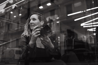 A young female photographer holding her Canon camera as she looks past her blond wavy hair, as the city lights reflect onto glass behind her.