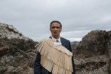 A Māori kaumātua dressed in a special cloak and wearing a bone carving around his neck, on top of a navy blue suit, standing in front of rocks on a cloudy day.