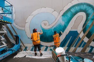 Two workers in high vis jackets painting a mural on the side of a building.