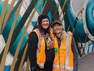 Two women in high vis jackets standing infront of a mural.
