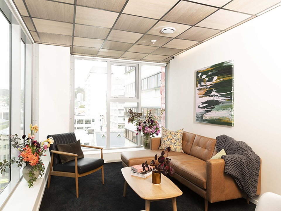 A lounge room with a brown leather couch on the right, a coffee table in front of it and a single chair. The couch over looks two large windows looking out to city buildings.