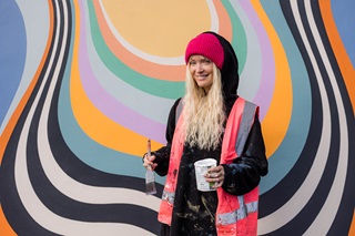 Woman wearing a pink high vis vest holding onto a paint brush and canister infront of a mural.