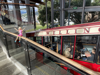 A barbie doll wearing a pink dress sitting on a ledge infront of the cable car.