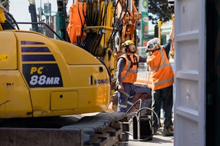 Two workers dressed in hard hats and high vis vests, talking next to a large yellow digger.