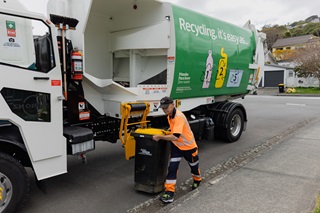 Person wearing a high vis orange shirt moving a yellow and black rubbish bin away from a recycling truck.