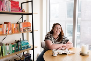 Woman wearing a striped tshirt sitting between the window and a bookshelf, with a book open on the table infront of her.