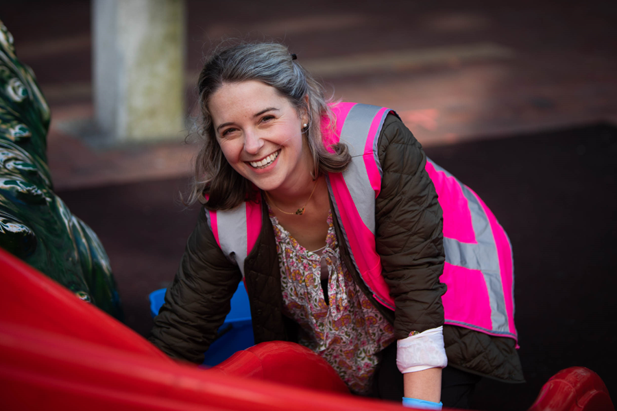 A young woman smiling while wearing a pink high-vis vest, as she scrubs a red plastic slide.