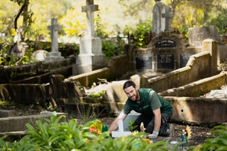 A young man looks at the camera as he crouches down on the dirt floor among grave stones in a cemetery. He is wearing work clothes and gloves for pulling weeds.