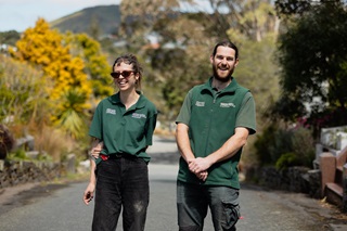 A young man and woman wearing green polo shirt uniforms smile as they stand on a concrete road, with autumn-coloured trees surrounding them.