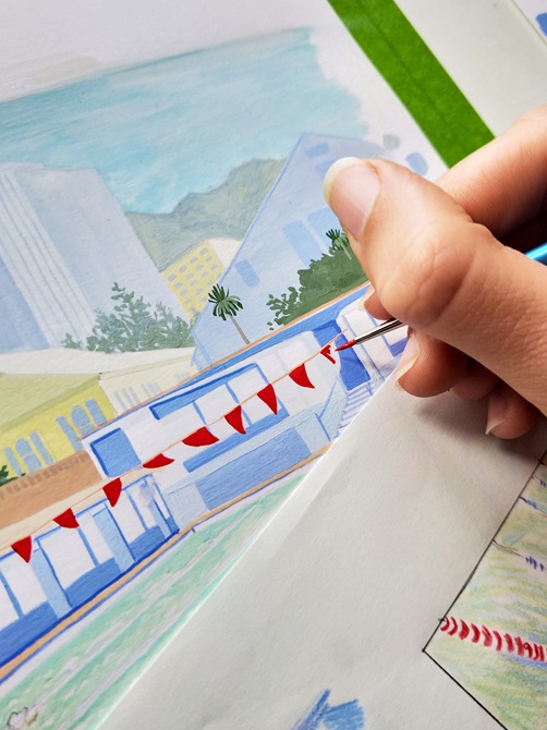 A hand using a small paintbrush to paint a watercolour painting of an outdoor pool.